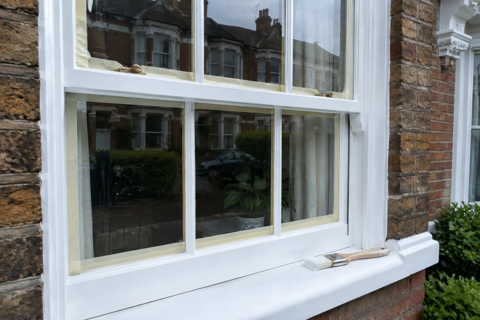White timber sash window on a brick house mid-repaint, with masking tape on the glass and a brush on the sill